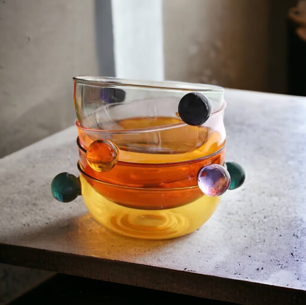 Four variations of a colorful borosilicate glass dessert bowl stacked one on top of the other. Each bowl features a distinct hue (transparent brown, smoky gray, pink, light yellow) and is equipped with two contrasting colored glass ball handles, creating a matching but vibrant set of serveware.