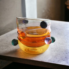 Four variations of a colorful borosilicate glass dessert bowl stacked one on top of the other. Each bowl features a distinct hue (transparent brown, smoky gray, pink, light yellow) and is equipped with two contrasting colored glass ball handles, creating a matching but vibrant set of serveware.